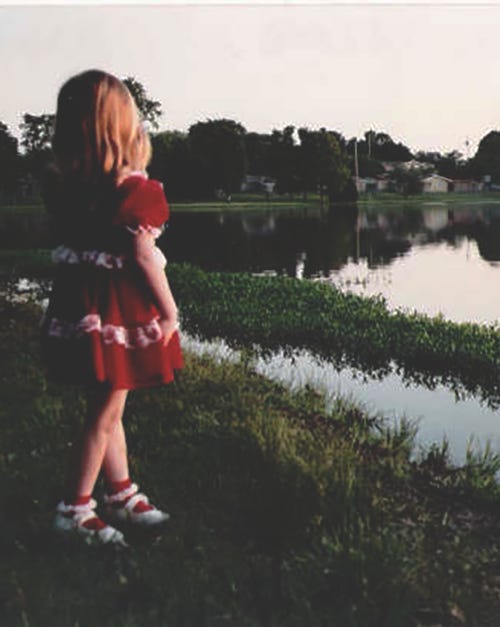 a young girl-child with shoulder-length blonde hair wearing a red dress stands in swampy area, looking at a body of water