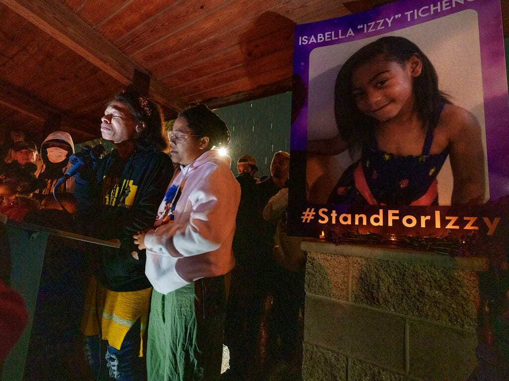 Two Black women stand at a podium. One of them is struggling to speak through her tears. They stand with others under a pavilion in the rain. On a wall next to them is a large poster with an image of a Black girl smiling for the camera. Isabella “Izzy” Tichenor. #StandForIzzy.