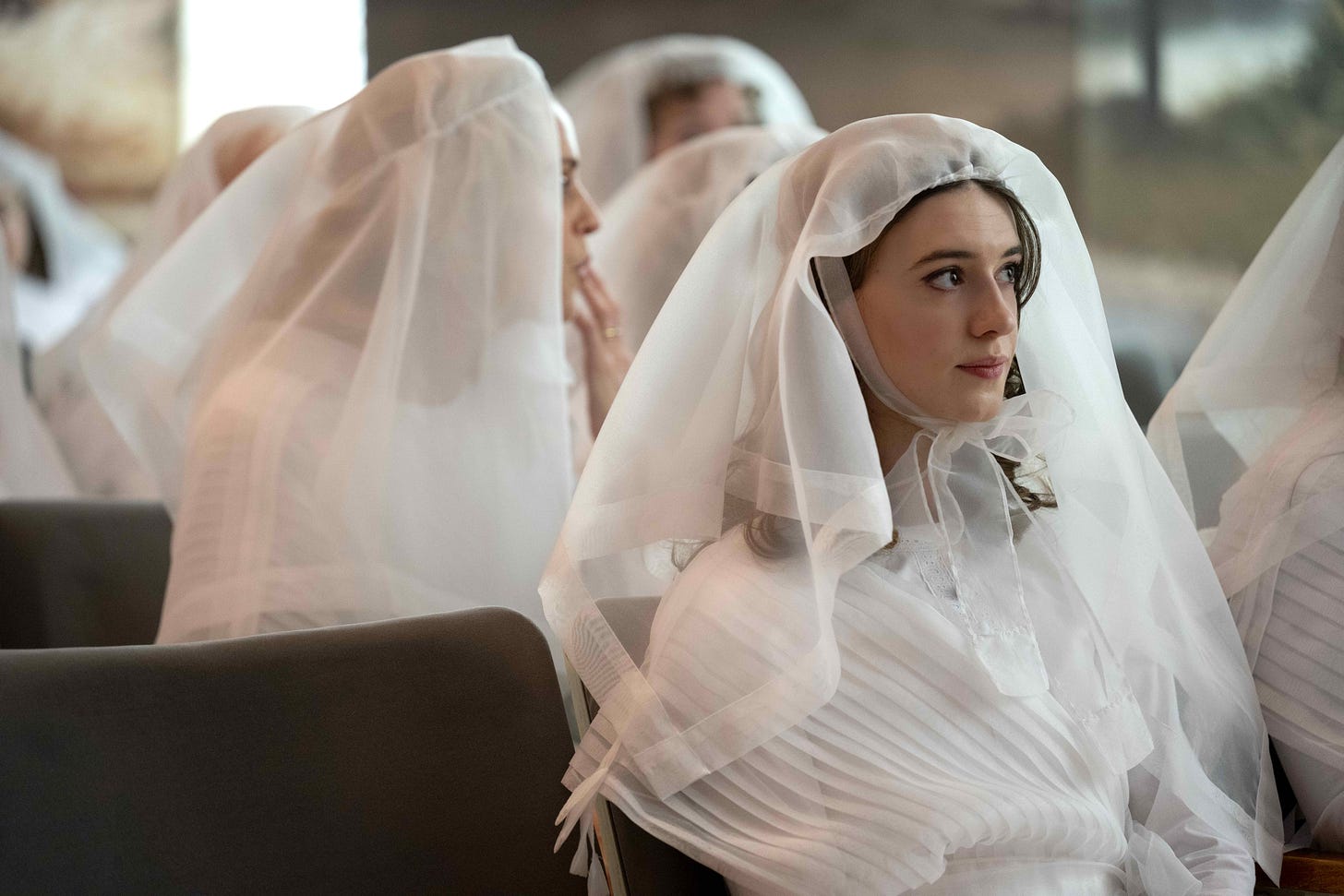 A young woman clad in the white robes and veil of the Mormon temple sits in sharp focus near the camera, while around her other similarly dressed women sit out of focus in other rows of seating.
