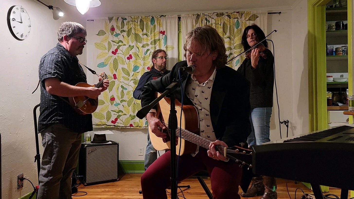 Musicians playing mandolin, accordion and violin surrounding a singer/ guitarist, in a living room