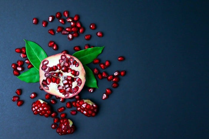 A pomegranate (cut in half, flesh up) is on a dark surface, surrounded by pomegranate seeds and a few bright green leaves