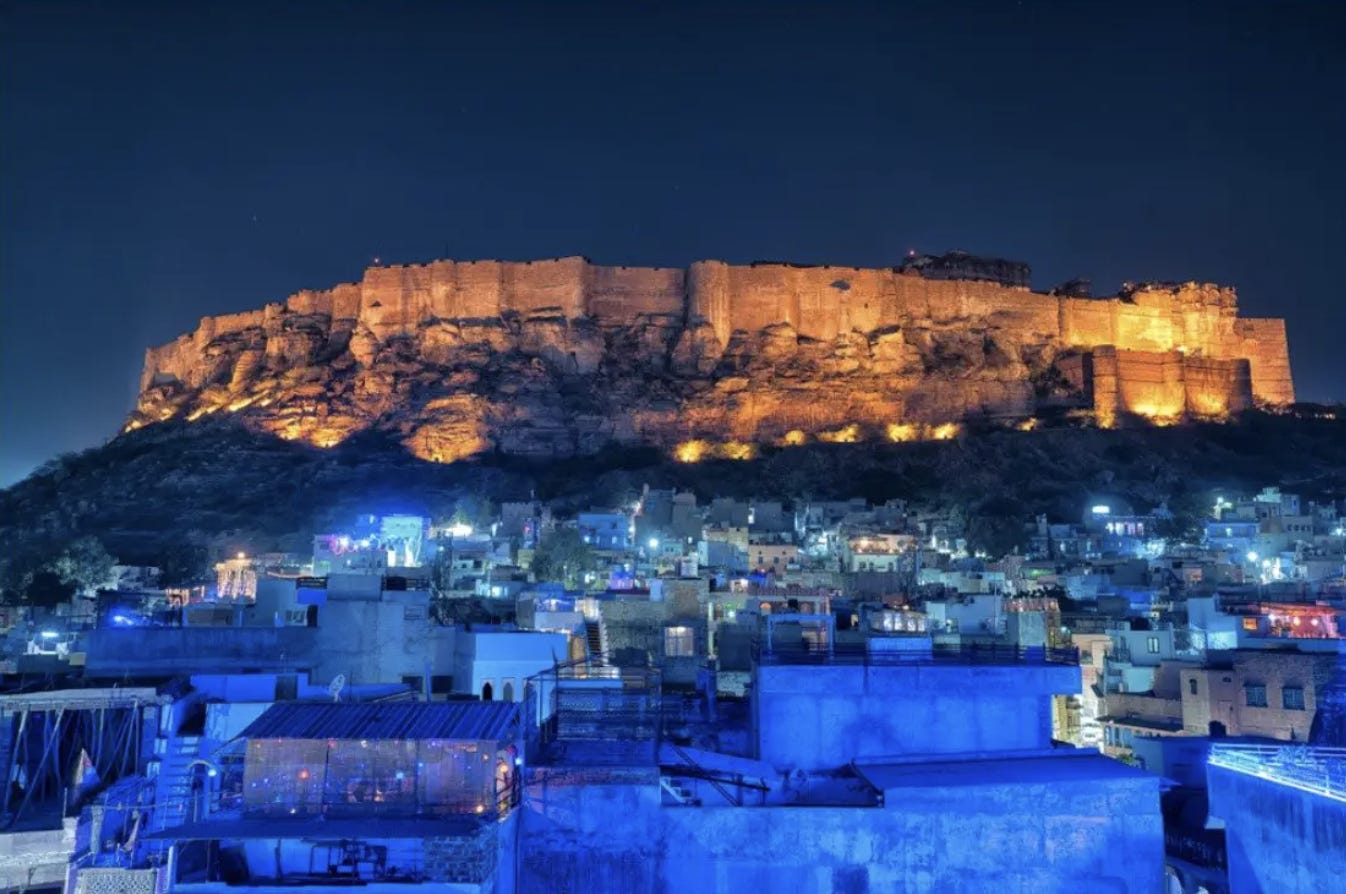 A city filled with blue buildings lit up in the night causing a blue glow. Above the city is a fort on a mountaintop lit up with golden light. The sky above is dark midnight blue.