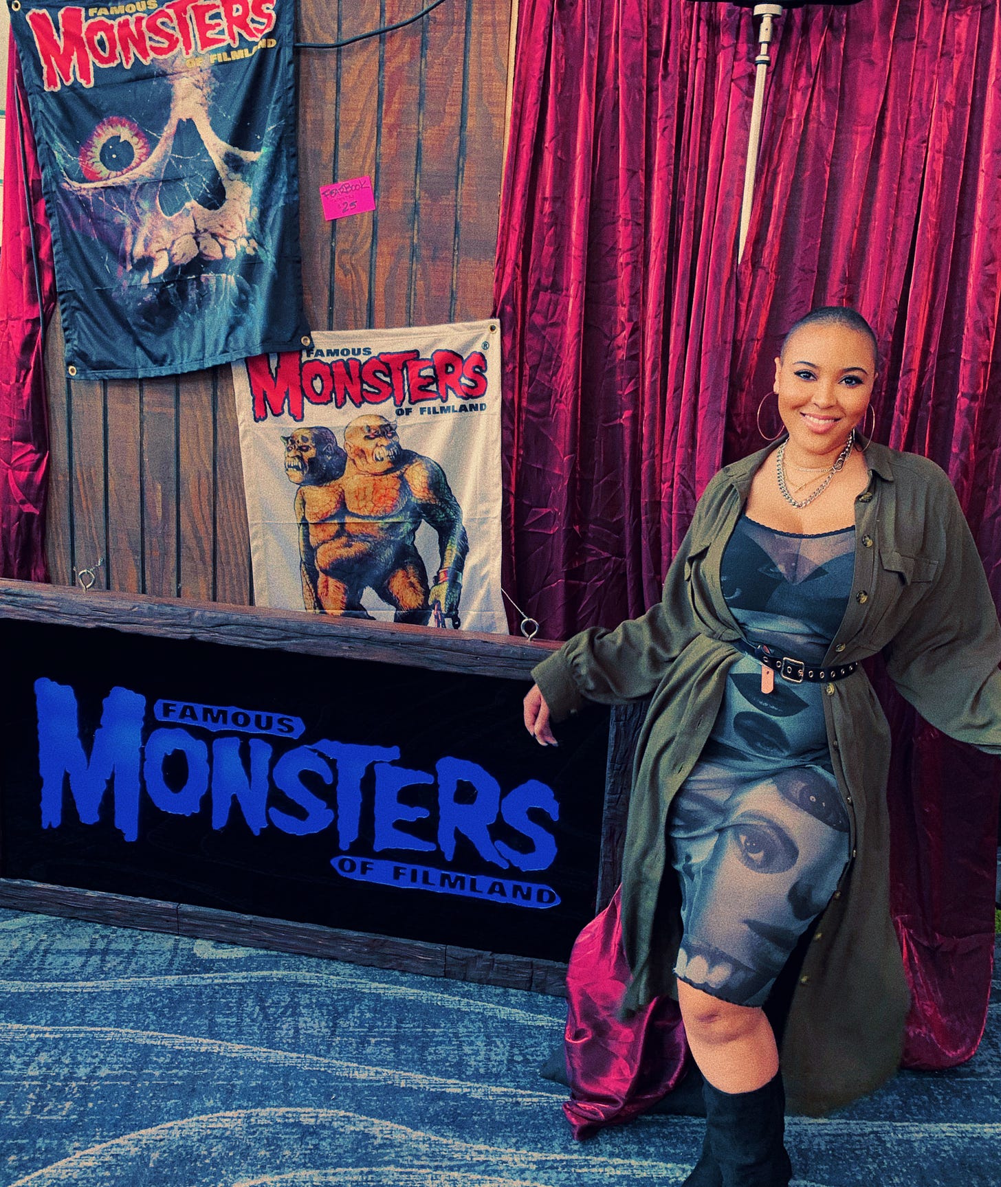 Beautiful bald woman (Lakyn, the author) standing in front of a backdrop for Famous Monsters magazine in a mesh dress with printed eyeballs and lips, layered gold and silver necklaces, a gold grommet belt and an olive maxi shirt dress. She's smiling and looks super hot.