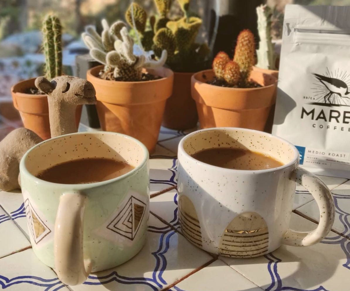 Two clay coffee mugs filled with coffee on a white tile table in front of small orange clay pots with succulents in them. Off the the right a white bag of Marea Coffee Medio Roast. To the left a clay camel is smiling.