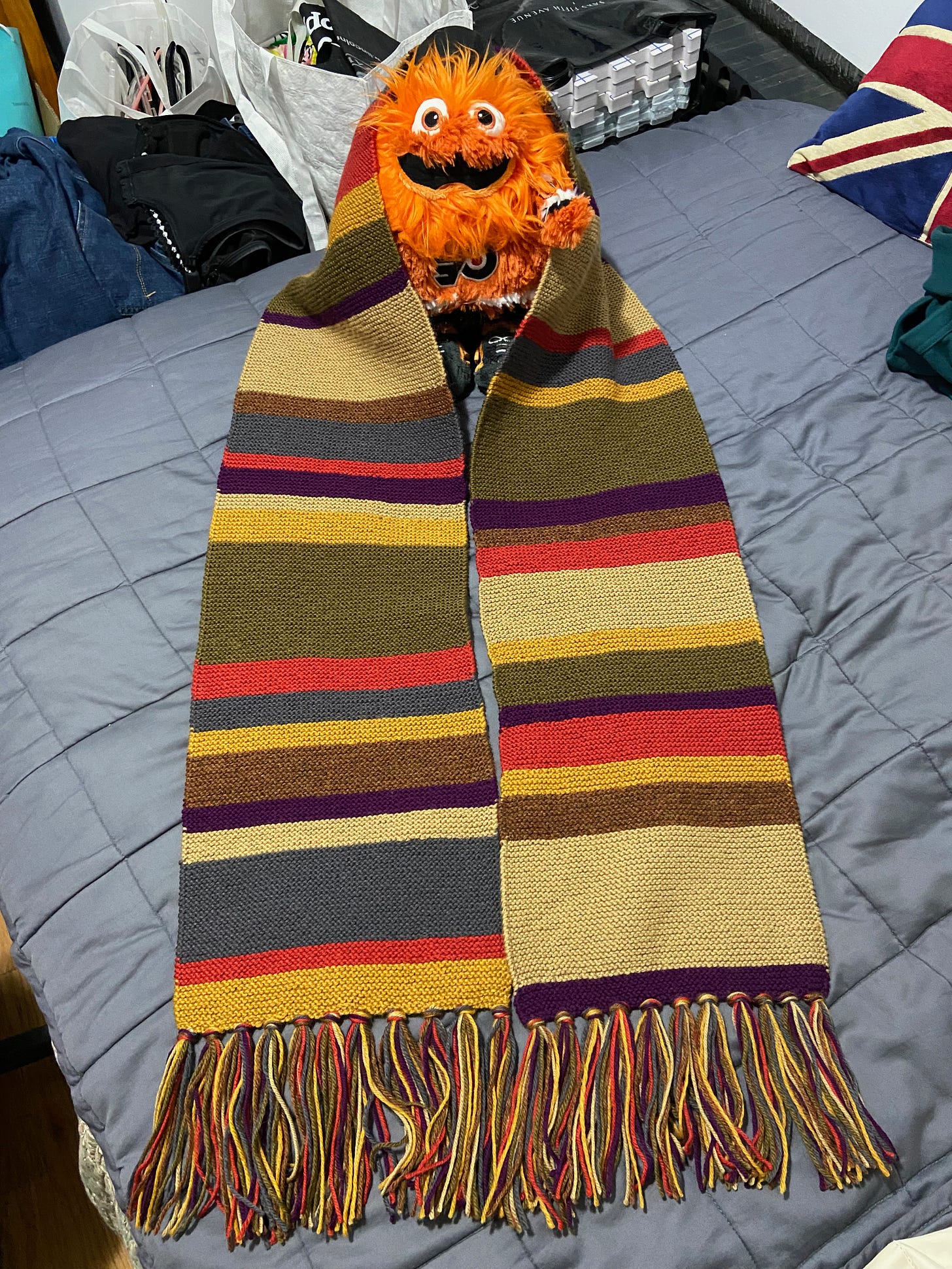 A fully knitted DOCTOR WHO scarf positioned around a miniature Gritty plush.