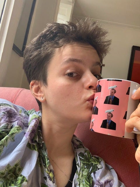 Rowan, with short brown hair and wearing a floral shirt, mimes kissing a pink mug with images of Niles, a man wearing a shirt, with a white cockatoo sat on his head