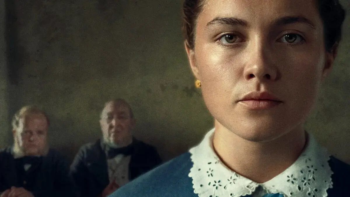 A still image from The Wonder. Florence Pugh in period dress fills the foreground, right. Two men sit, out of focus over her right shoulder.