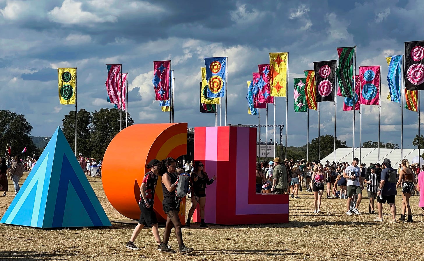Colorful large block letters, A with shades of blue stripes, C in shades of orange stripes, and L with red and pink stripes, sit in the liddle of Zilker park. Colorful flags wave against a stormy clouds as people mingle around the park. A music stage sits in the background.