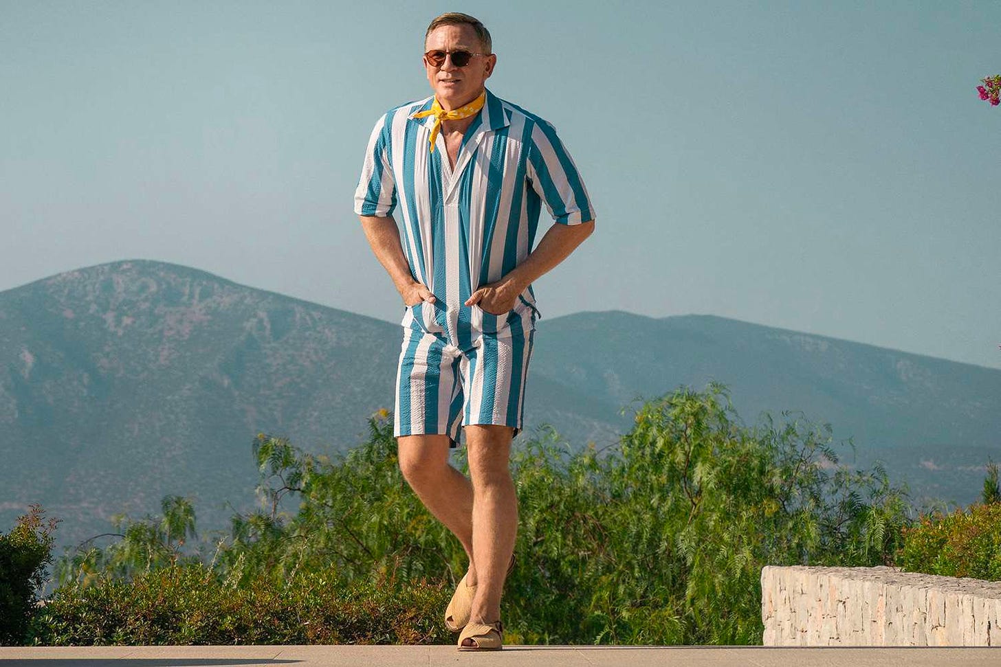 Daniel Craig as Benoit Blanc wearing a white and blue striped romper by the pool.