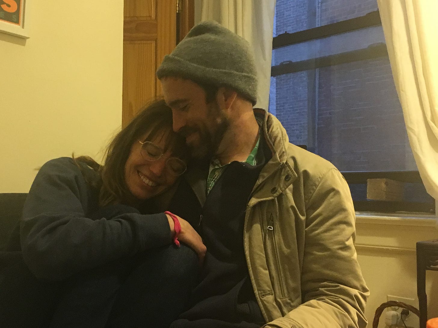 A white woman with brown hair snuggles into a white man with a beard and a hat. Both look to be in their thirties. 