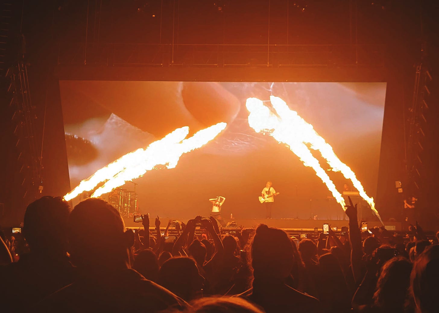 a concert, halsey is on stage singing on their knees, with an image of skin behind them and streaks of pyrotechnic fire on both sides of the stage.