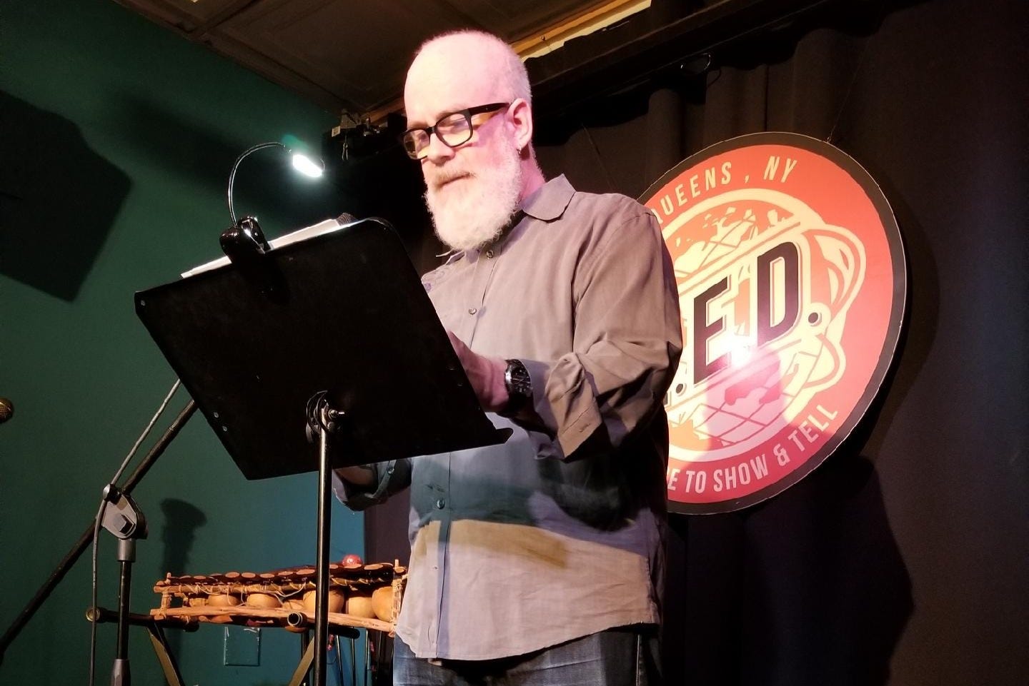 A gray-bearded, bespectacled man stands in front of a microphone reading from a sheaf of papers on a music stand, apparently on a very small stage. He is spotlight, while behind him on a black curtain hangs the logo for the Q.E.D. comedy club.
