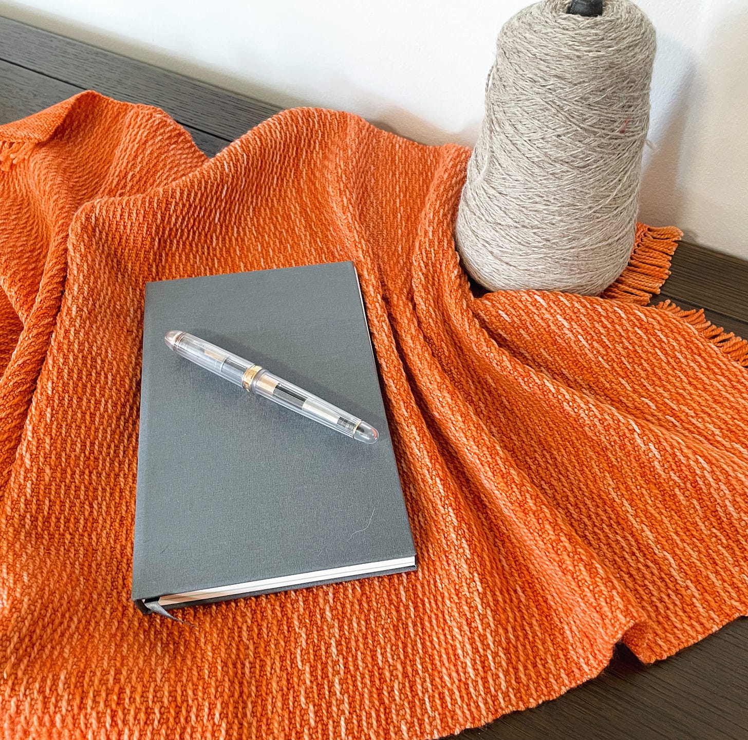 A pen on a gray notebook on an orange cloth on a brown surface. A cone of yarn nearby.