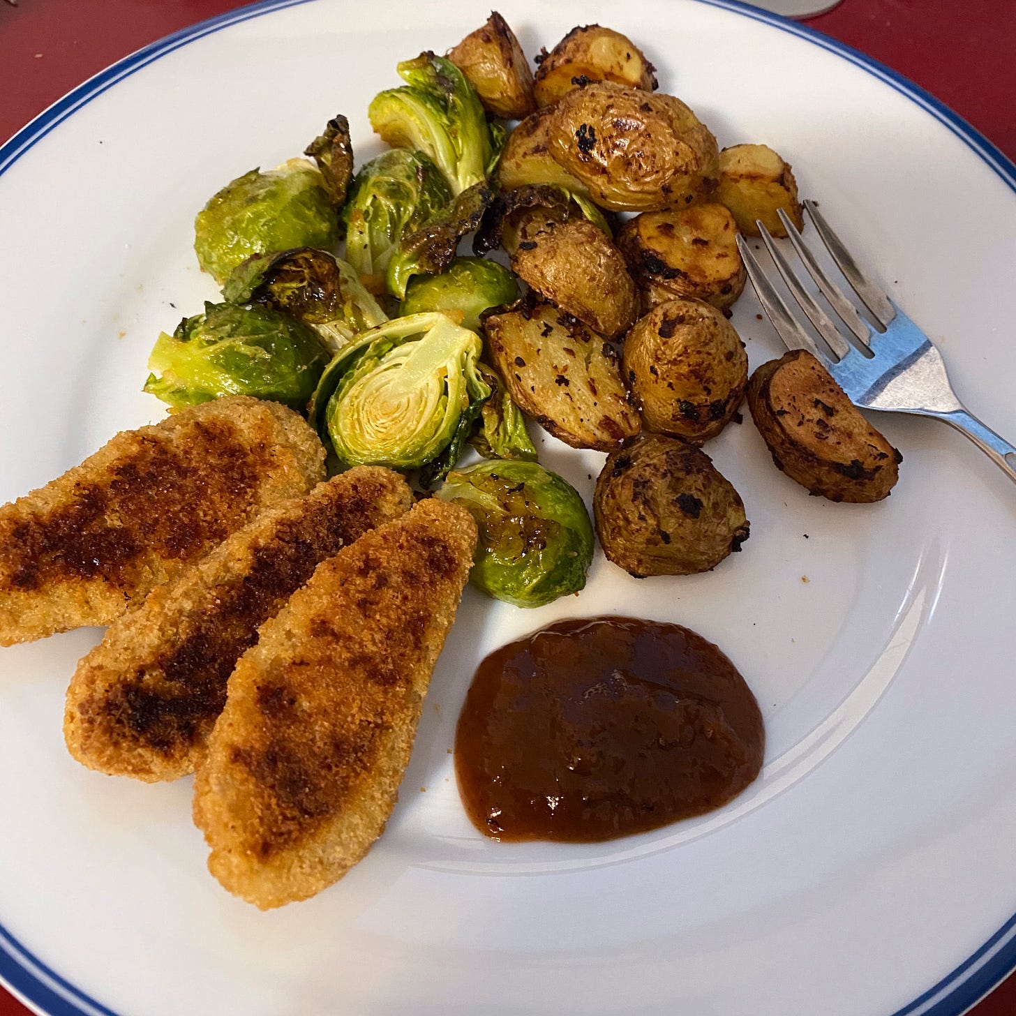 On a white dinner plate with a blue rim, three toasted vegan nuggets next to a splotch of dark brown plum sauce. Behind the nuggets are piles of roasted potatoes and brussels sprouts. A fork rests at the right edge of the plate.