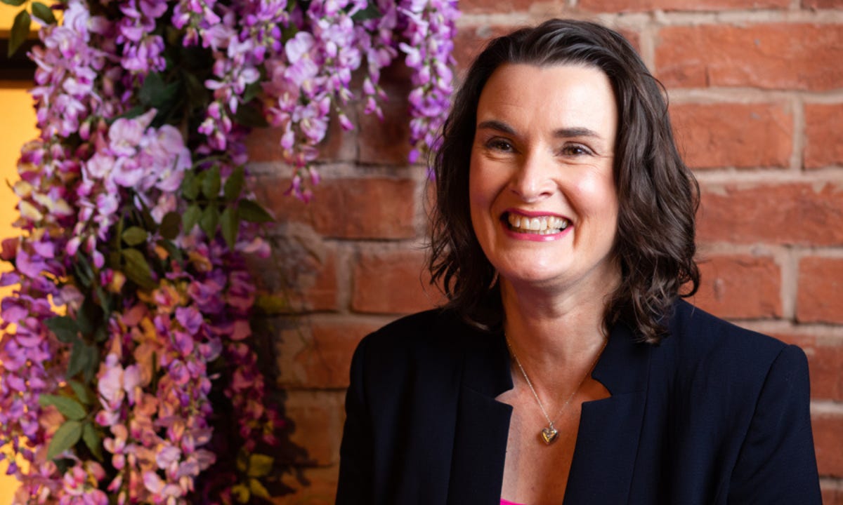 Photo of Lucinda smiling, standing in front of a wall with wisteria flowers behind her