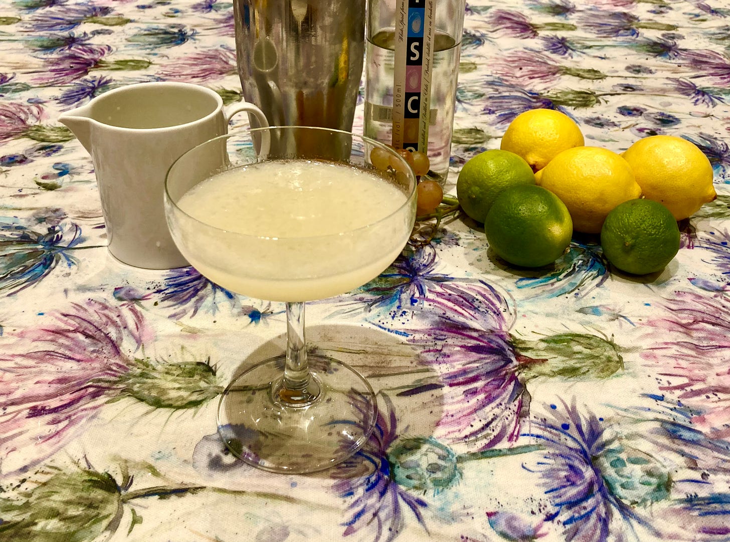 A Pisco Sour in a coupe glass, on a table with a shaker, jug of citrus juice, bottle of pisco, and some citrus fruit as decoration.