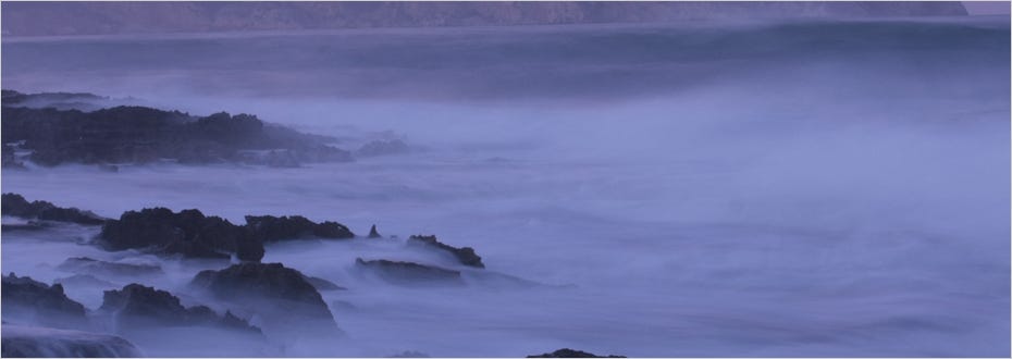 A heavy mist covers rocks on a turbulent shore at twilight.