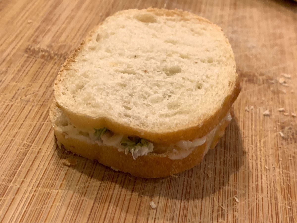 A small sandwich of sliced baguette, butter, and a couple of visible clover blossoms sits on a wooden cutting board with crumbs behind it. 