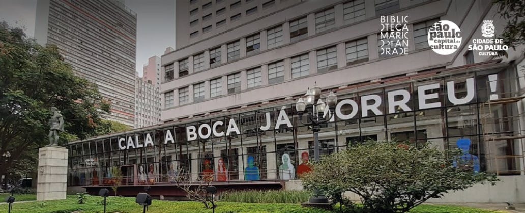 Varanda do prédio da Biblioteca mário de Andrade cercado de vidro com as palavras CALA BOCA JA MORREU escritas em letras brancas enormes.