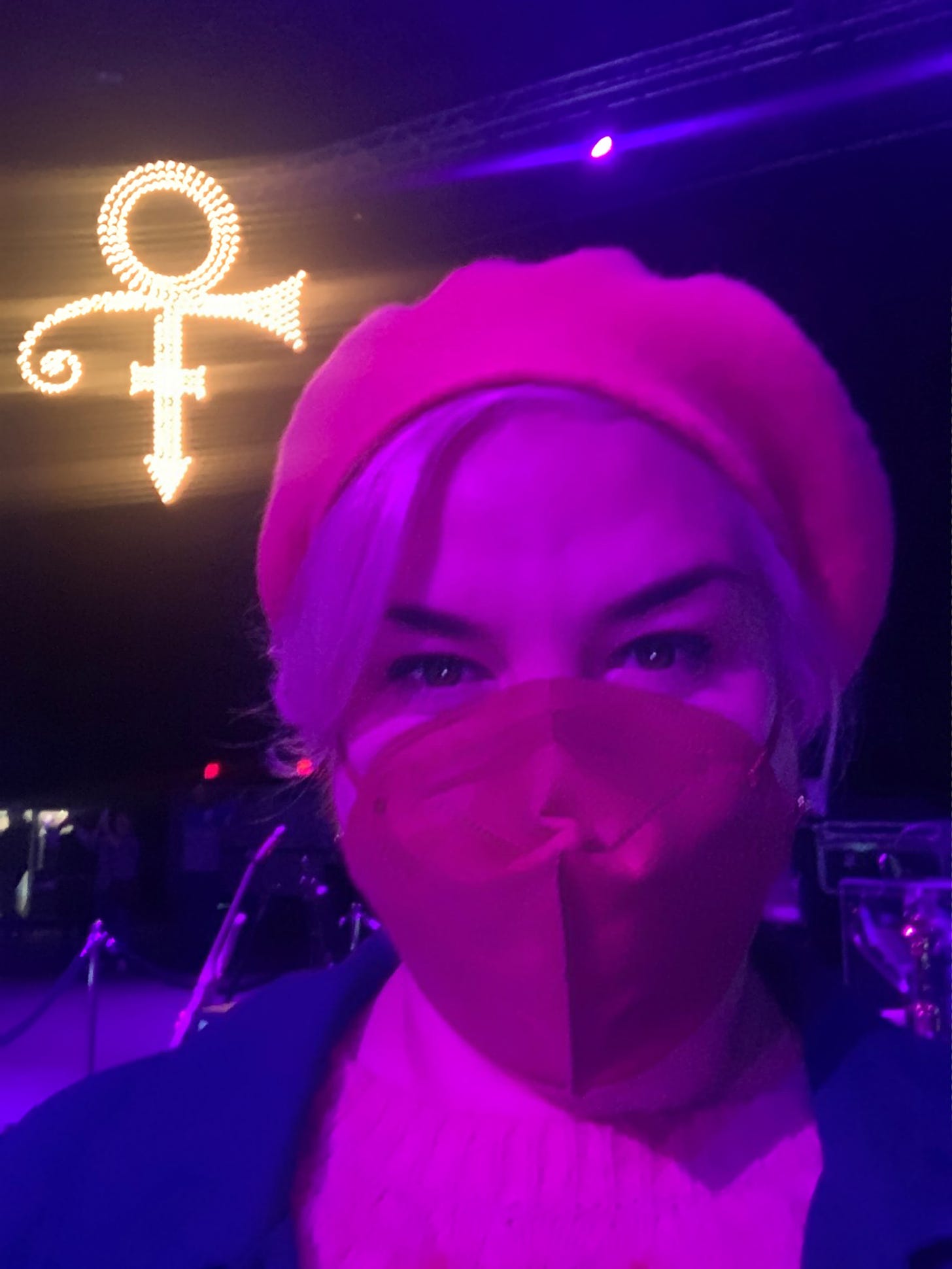 Caissie in a beret and face mask stands on a darkened soundstage at Paisley Park in front of an illuminated Love Symbol, Prince's signature.