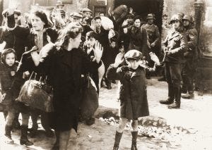 Photo showing women and children with their hands raised in front of Nazi soldiers with guns in the Warsaw ghetto during World War II.