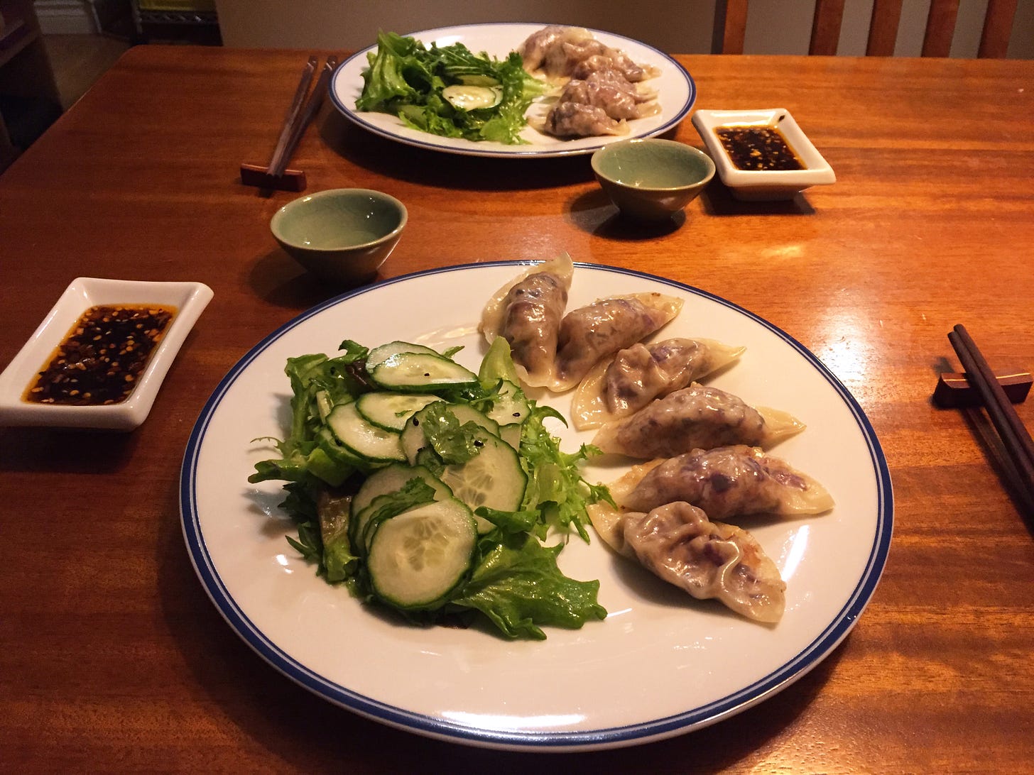 Two plates with rows of 6 dumplings and a pile of cucumber and endive salad with black sesame seeds. Next to the plates are chopsticks, small cups of rice wine, and flat dishes of sauce with chili flakes visible.
