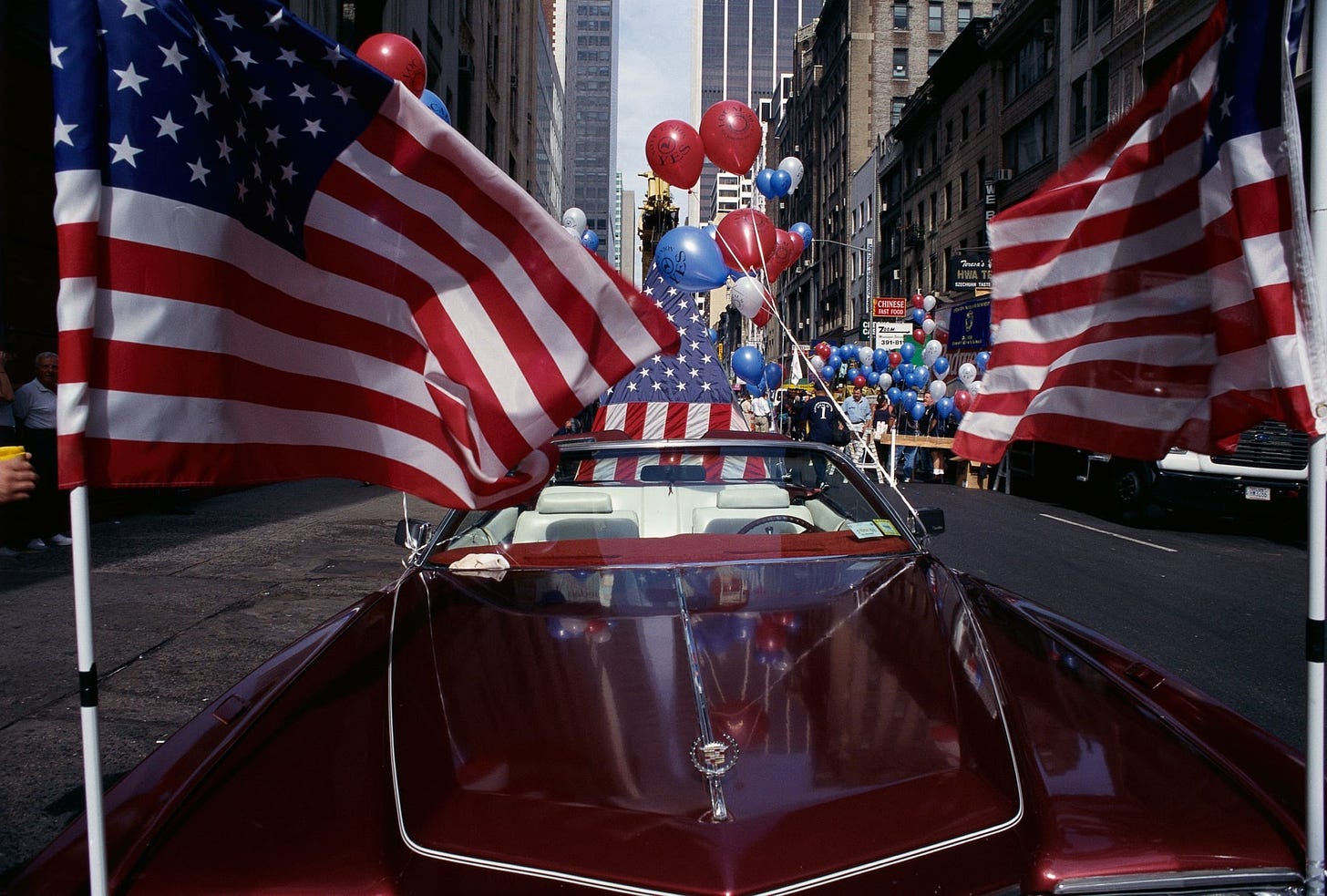 red white and blue balloons and many american flags attached to a red cadillac on the streets of new york city