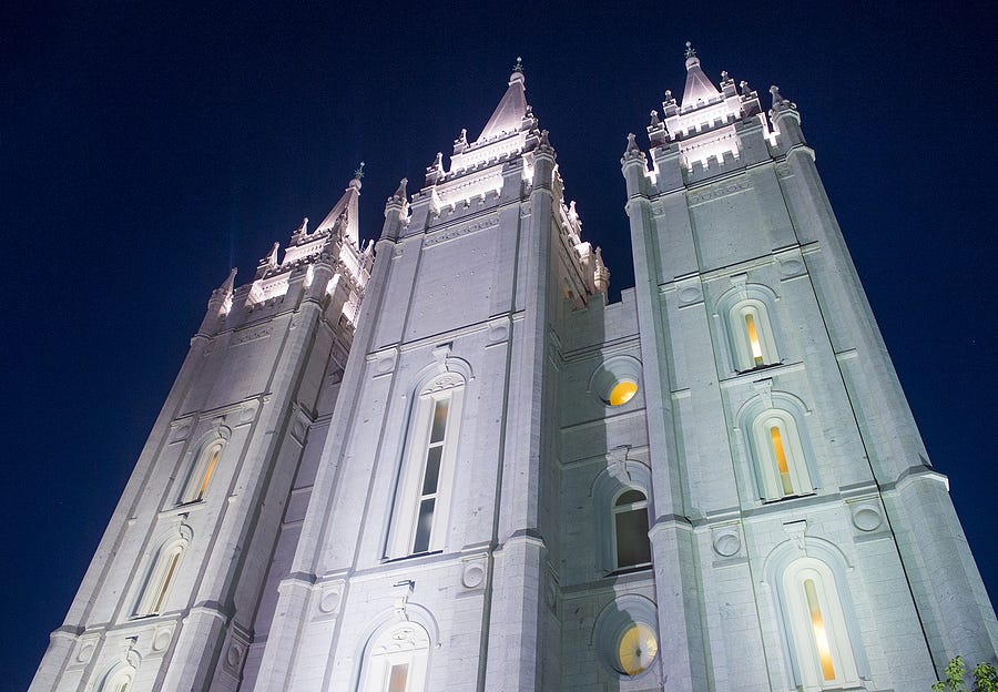 A view looking up at the illuminated gray granite facade of the Latter-day Saint (Mormon) Temple in Salt Lake City, Utah, with three spires visible in front of a purplish night sky.