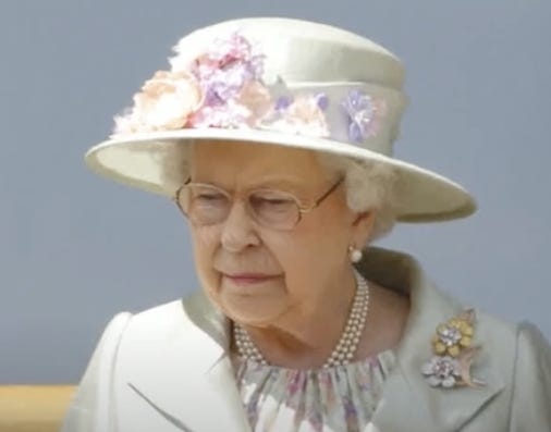 A picture of Queen Elizabeth during the day time looking outwards. Queen Elizabeth is in a suit coat with a pastel flower print top underneath. She’s wearing a pearl necklace and a giant broach on her left shoulder, and a matching hat with pastel flowers on it. She is not smiling. There is a grey background behind her.