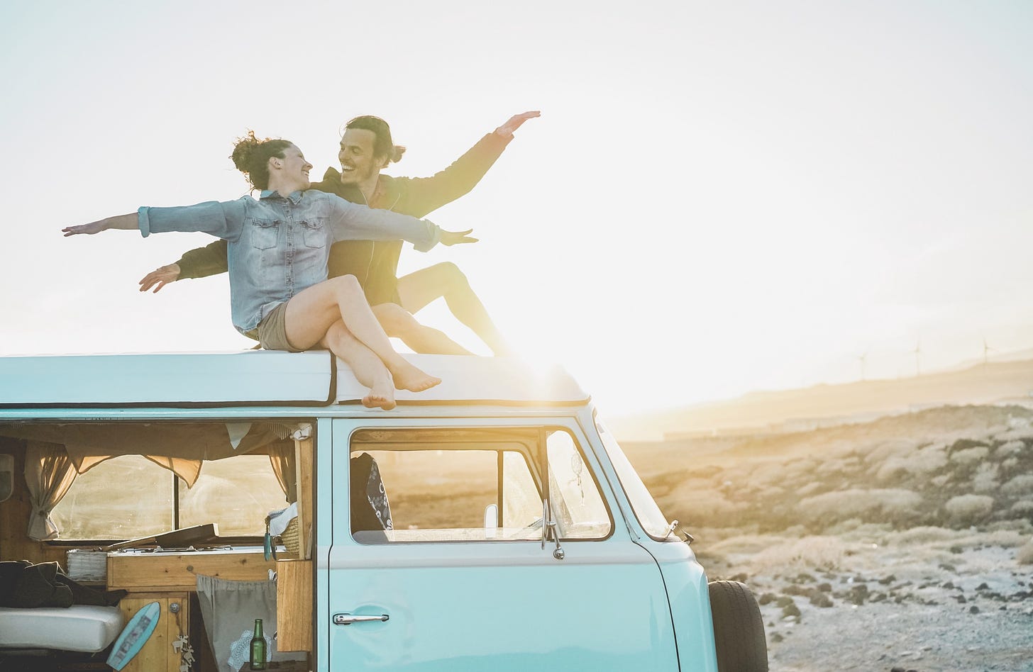 A silly couple sits on top of their van at sunset with their arms spread wide.