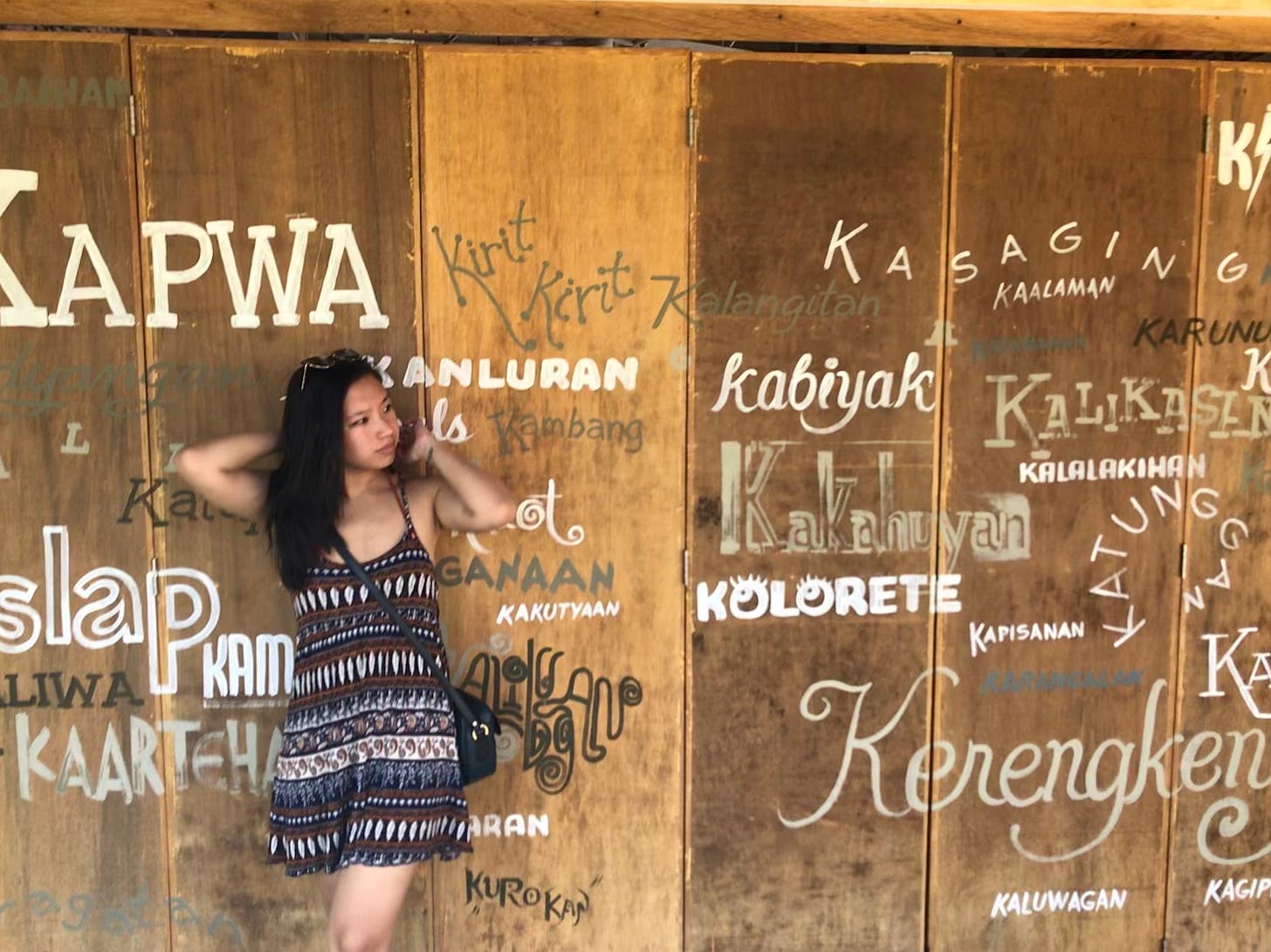 Justine in a summer dress standing against a wooden wall with Tagalog words painted on it. 