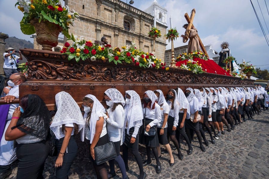 Dozens of women walk on a cobblestone street, supporting a large wooden float with a statue of Jesus Christ on top.