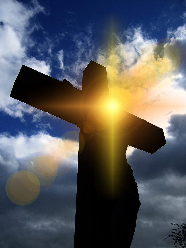 Against a bright blue background with fluffy white clouds, a silhouette of a man, likely meant to represent Jesus, is hung upon a thick wooden cross in shadows. None of his features are evident, however, a bright yellow ray of sunshine shoots past him, directly at the camera.