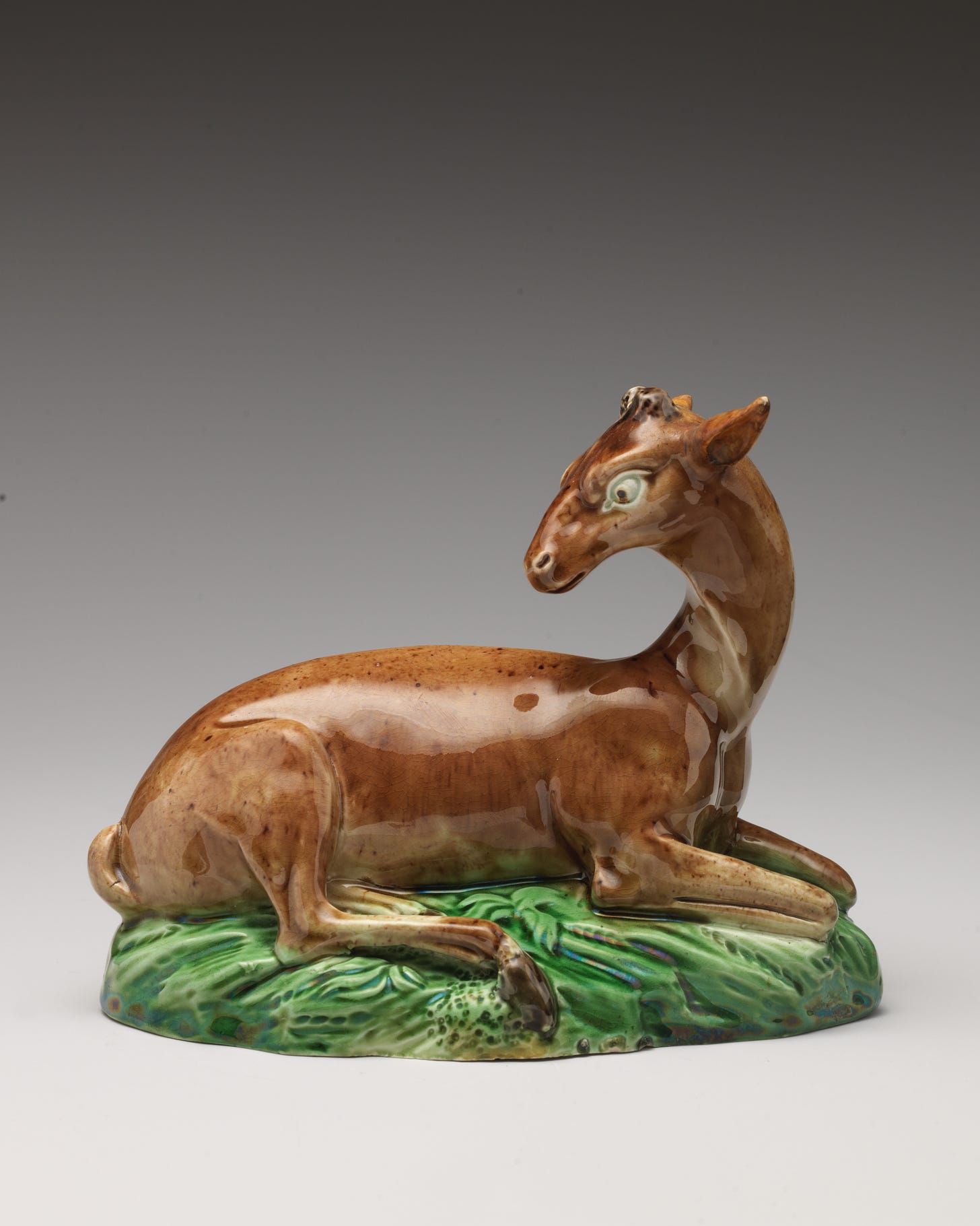 Ceramic brown doe with large eye, arching neck towards back, sits atop green ceramic grass.