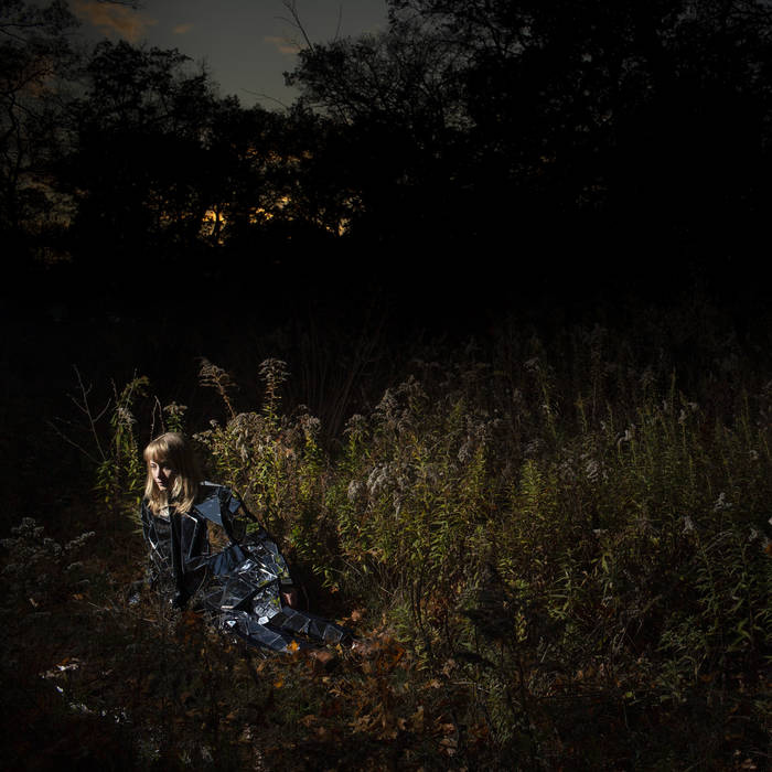 The album cover of Ignorance, depicting a dusk scene with a blonde woman, Tamara Lindeman, lying on her side in a forested area while wearing a reflective suit