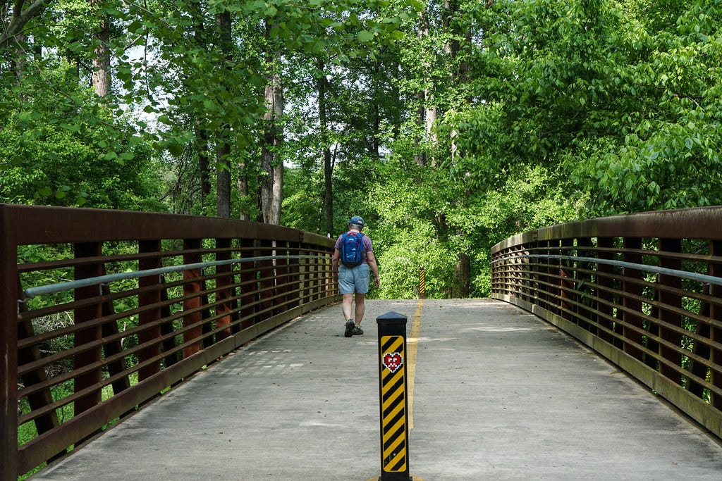 A man walking over a bridge on the Atalanta Beltline. There are a lot of green trees ahead of him as he walks away from the camera