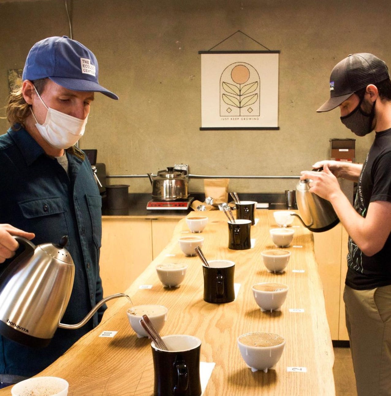 Two men on opposite sides of a long wooden table pour water out of goose-neck kettles into coffee cupping bowls (like small soup bowls)