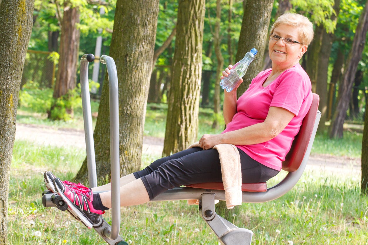 Older white, smiling woman on a piece of outdoor excercise equipment in a park. She's holding a bottle of water.