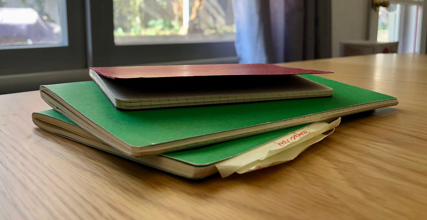 a messy stack of three worn notebooks on a wood table with sunny windows behind