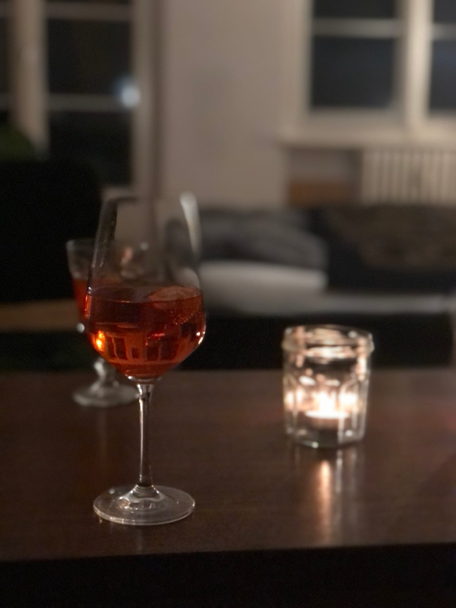 An evening scene. A glass of pinkish liquid set on a low table. Just behind it, another differently shaped glass containing the same pink liquid. Beside this, a jar with a candle lit inside it. 