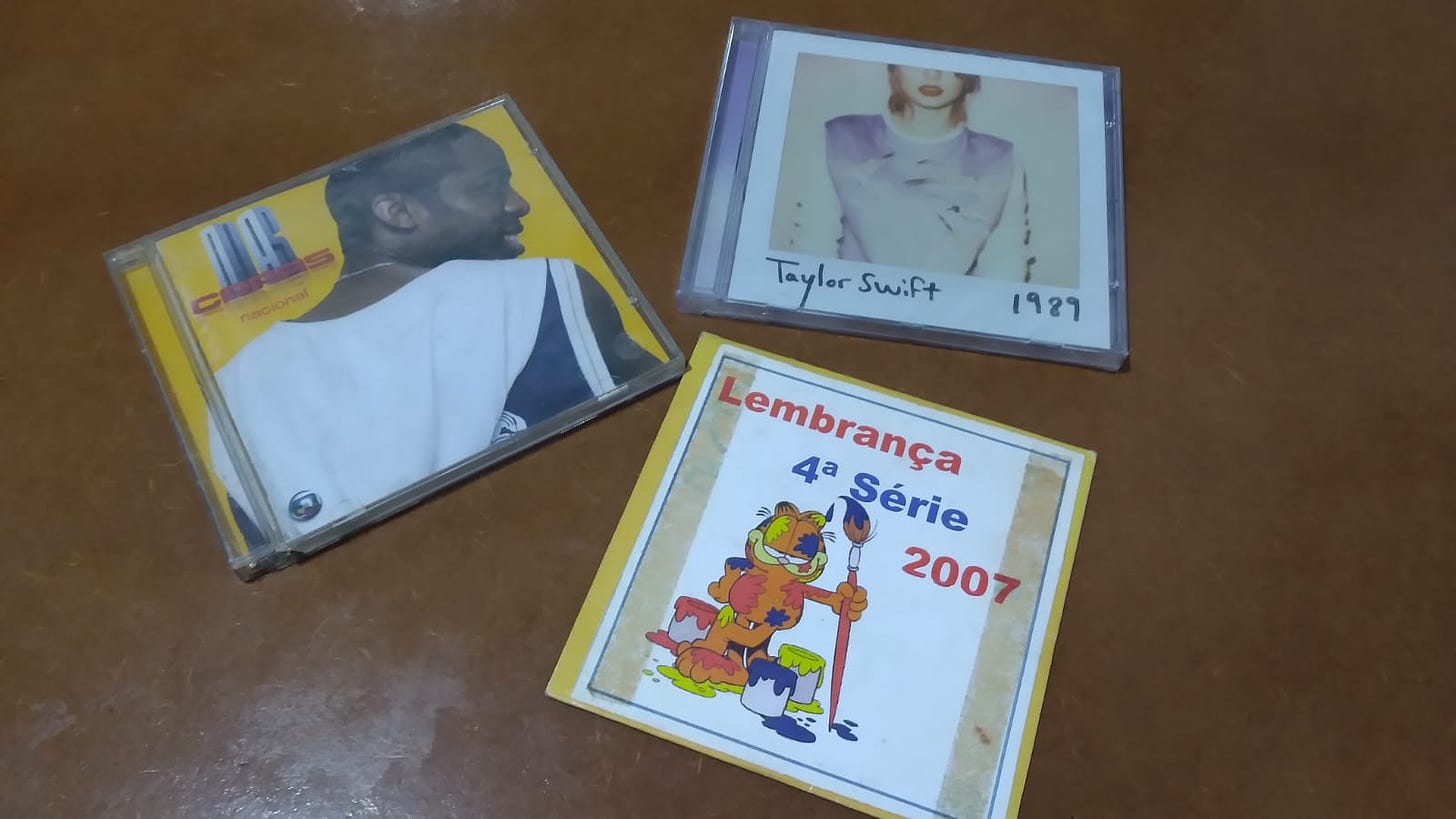 Foto com os CDs "Duas Caras Nacional" e "1989", de Taylor Swift, em cima de uma mesa de madeira escura, junto de uma capa de CD de papel, com uma etiqueta impressa em papel com os dizeres "Lembrança 4ª série 2007" e uma ilustração do Garfield, com baldes de tinta e um pincel, e coberto de manchas de tinta.