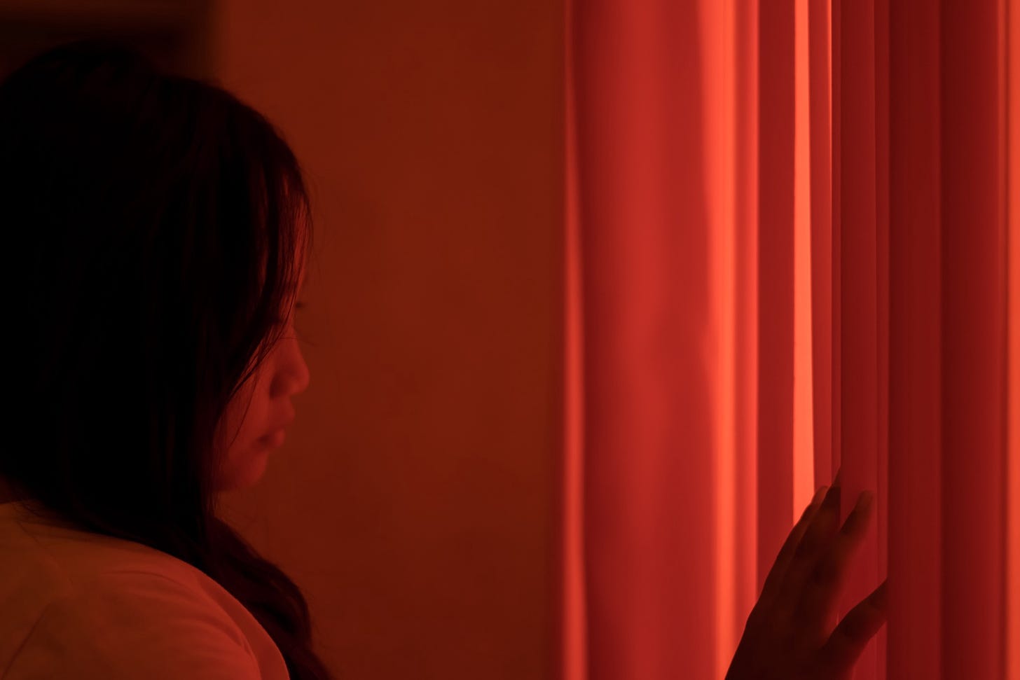 A young woman bathed in red light stares through a curtain.