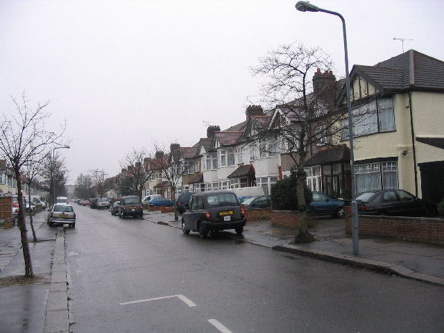 a classic suburban British street. Wide road, cars parked on the pavement, and a long line of terraced and semi-detached housing. and its grey and wet
