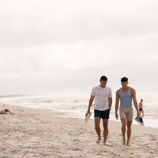 Two men in shorts and t shirts taking an intimate walk on a Fire Island beach, with the caption: ‘Fire Island’ writer and creator Joel Kim Booster (right) plays Noah, who finds love with Will (Conrad Ricamora, left) because, well, opposites attract, in Jane Austen and on Fire Island. Photo: Courtesy Searchlight Pictures