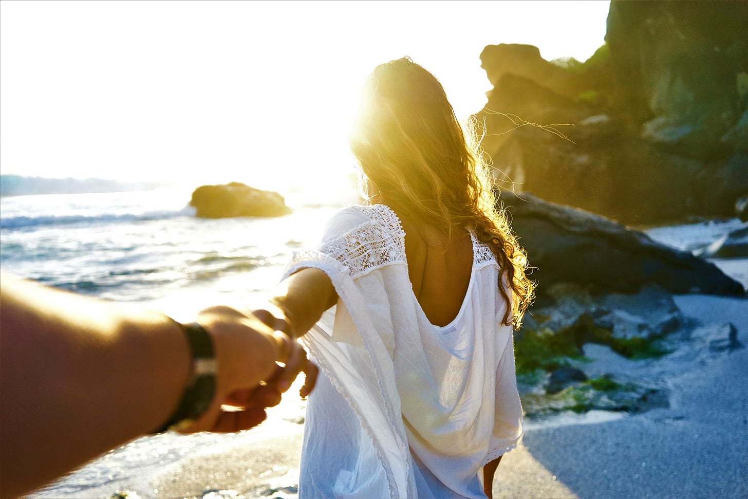 back view of woman wearing white shirt walking on beach and holding a man's hand