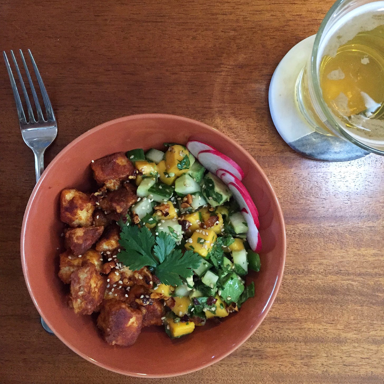 An orange bowl full of pieces of tofu in a red curry coating, and a salad of cucumber, mango, and avocado pieces. Cilantro and sesame seeds are on top, and a sliced radish is at the side of the bowl. Next to it on a coaster is a glass of lager.