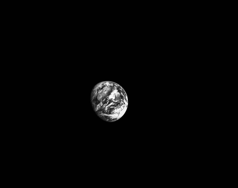A black-and-white image of the Earth from space. The lower left edge of Earth is in shadow, and the background is black and starless. White clouds are visible on the surface, with the grey shape of the African continent visible underneath.