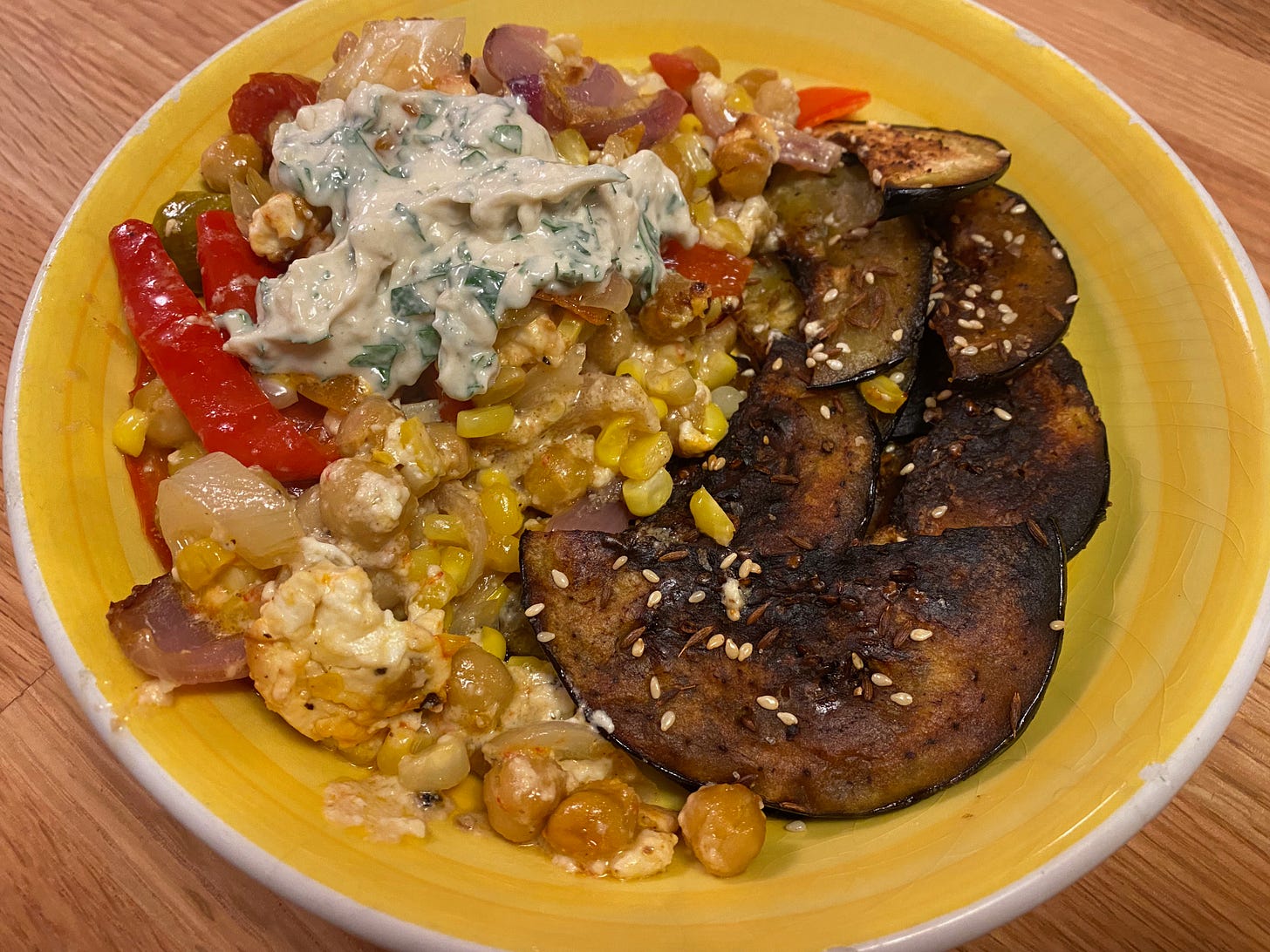 A wide yellow ceramic bowl filled with roasted corn, peppers, and chickpeas on one side and crispy half-moon eggplant slices on the other. The veggies are topped with a dollop of creamy yogurt tahini sauce flecked with parsley.