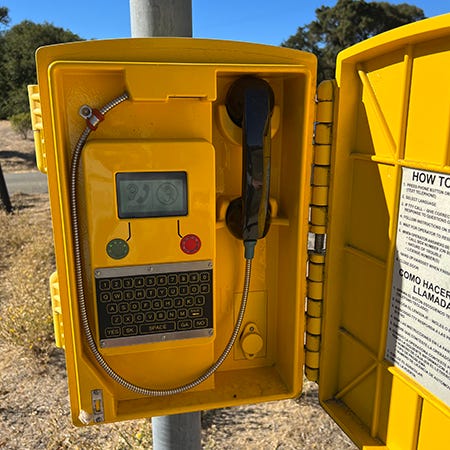 A picture of a California highway emergency call box, which is an open yellow case affixed to a pole with a black telephone receiver, a green button for audio, a red button for now audio, and a QWERTY keypad.