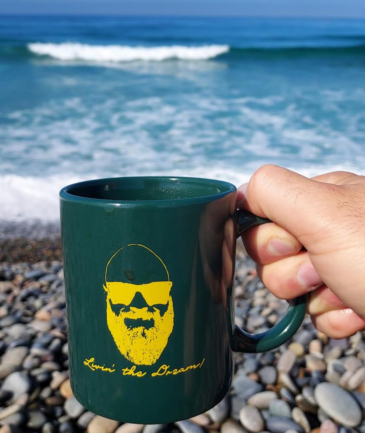 Close-up of a green coffee mug being held on the right of the frame up in front of a blurred wave crashing on beach rocks. The mug has a gold silhouette of a bearded man's head wearing sunglasses, and in text, it says, "Livin' the Dream."